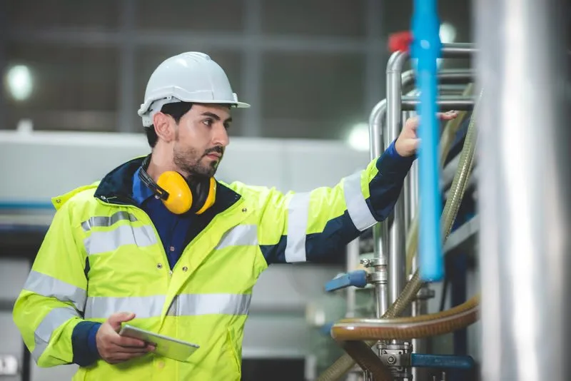 Operator with helmet, ear protection and tablet inspecting pipes in a process plant