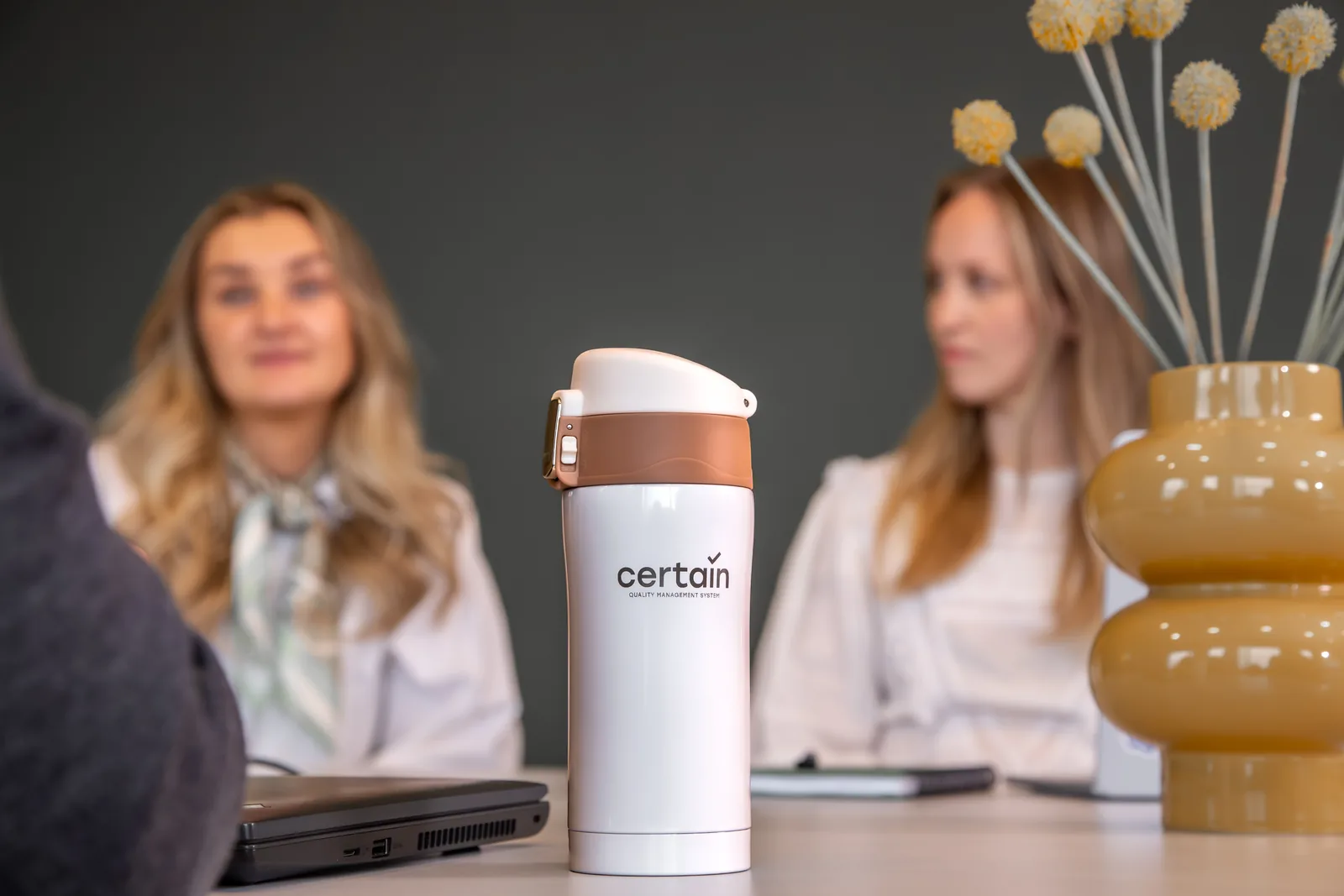 A Certain-branded travel mug on a meeting table, with two colleagues in the background